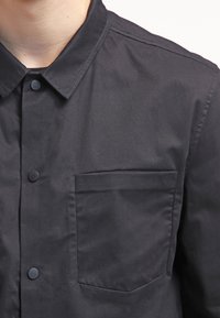 Mies, joka käyttää mustaa nappipaitaa, jossa on kärjellinen kaulus ja vasemmalla puolella rinta-tasku yksinkertaisen taustan edessä.