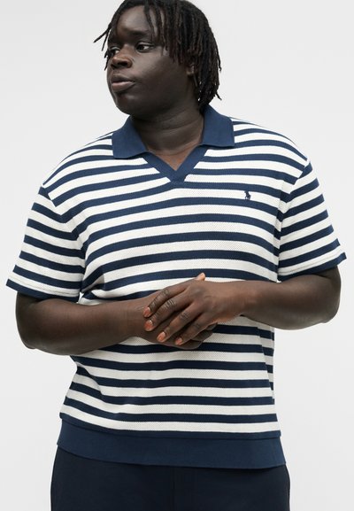 Bărbat cu pielea închisă la culoare și dreadlocks, purtând un tricou polo cu mânecă scurtă, în dungi albastre și albe, privind în parte cu mâinile împreunate.