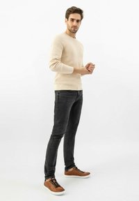 Beige pletený svetr, černé slim-fit džíny a hnědé tenisky s bílou podrážkou. Oblečení má ležérní, moderní design a přiléhavý siluetu.