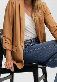 Světle hnědý blejzr s vyhrnutými rukávy, doplněný bílým topem a tmavě modrými džínami. Má volný střih a měkkou, hladkou texturu látky.