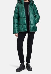 Zelená péřová bunda s kapucí, která má prošívaný design a lesklý povrch. Spárována s černými kalhotami a �černými robustními botami.