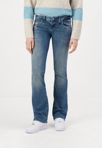Faded blue denim flared jeans with a five-pocket design, subtle distressing, and button closure. Worn with white sneakers.