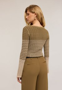 Pulover cu maneci lungi, având un model în zigzag în tonuri pământii de verde, bej și crem, combinat cu pantaloni maro cu talie înaltă.