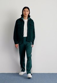 Zelena flis jakna z gumbnim zapiranjem, z dvema sprednjima žepoma; v kombinaciji z zelenimi trenirka hlačami z belimi stranskimi črtami in belimi supergami.