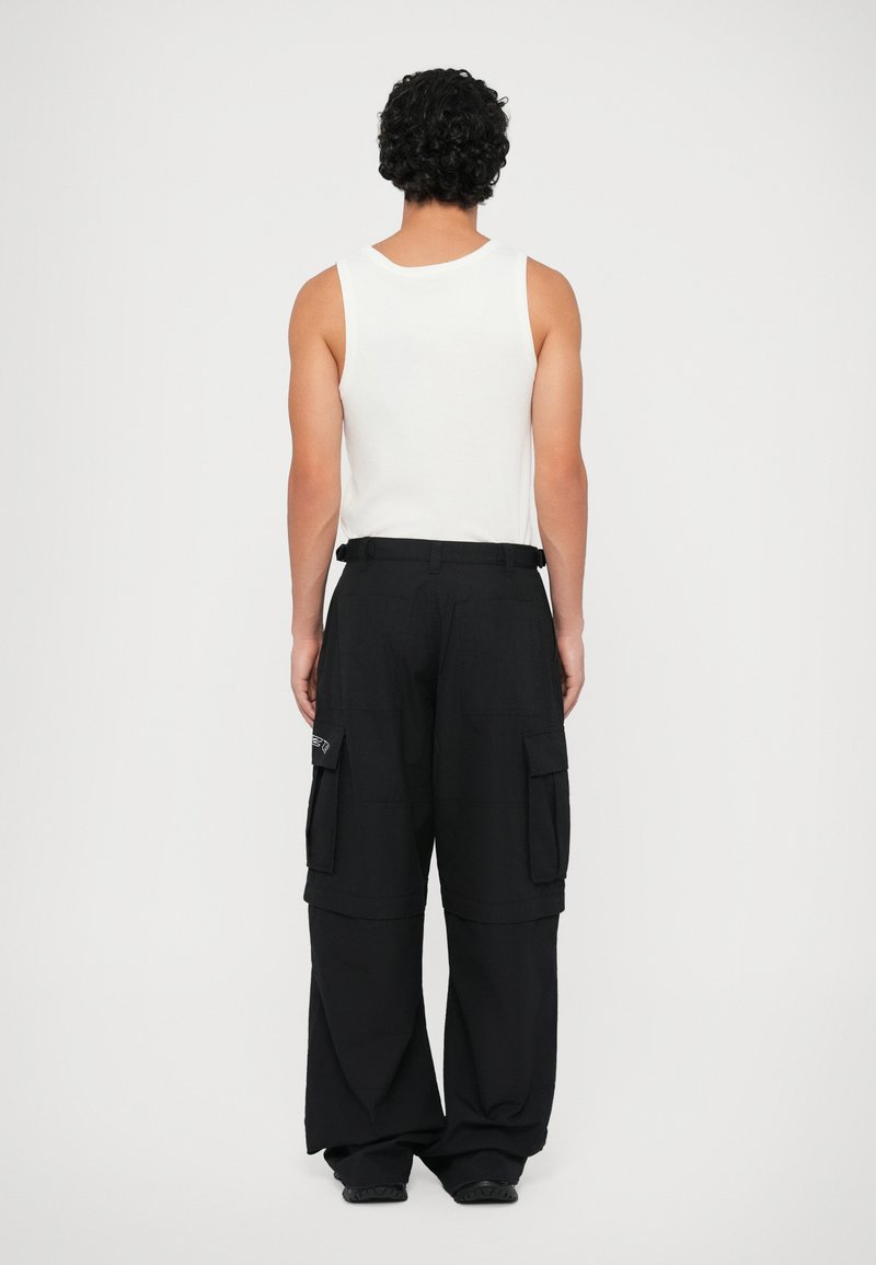 Schwarze Cargo-Hosen mit lockerer Passform, ausgestattet mit zwei Seitentaschen und einem Logo-Patch, kombiniert mit einem weißen ärmellosen Oberteil.