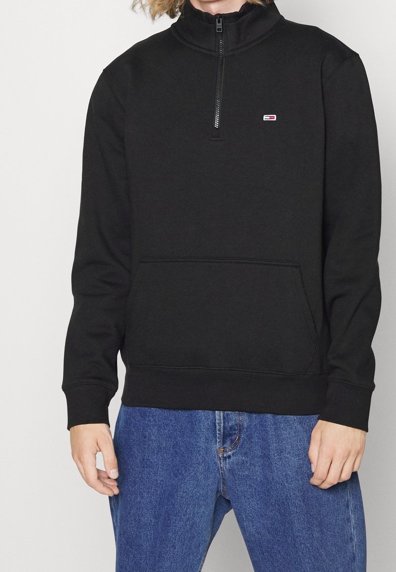 Personne portant un sweat-shirt noir à fermeture éclair quart avec un petit logo sur la poitrine et un jean bleu, debout devant un fond blanc uni.