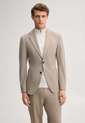 Junger Mann, der einen beigen Zweiknopfblazer und passende Hose über einem weißen Zip-Pullover trägt, steht vor einem schlichten hellen Hintergrund.