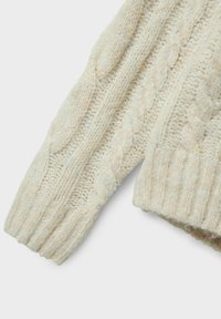 Manica di un maglione beige lavorato a maglia con motivo a trecce, polsino a coste e texture morbida, con variazioni nello spessore del filato per un interesse visivo.
