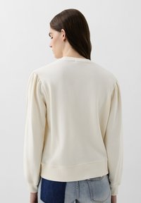 Cremefarvet sweatshirt med puffede ærmer, ribbede manchetter og kant samt en rund halsudskæring. Båret over blå denim med varierende teksturer.
