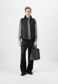 Zwarte gewatteerde vest over een grijze trui, gecombineerd met zwarte wijde broek. Houdt een gestructureerde zwarte leren tote bag vast. Minimalistische stijl.