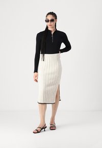 Svart langermet topp med glidelås i halsen, kombinert med en ribbet beige skjørt med sidesplitt og svart trim. Svart strappy hæler.