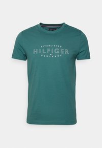 Teal-kankaista t-paita, jossa on pyöreä kaula-aukko, lyhyet hihat ja valkoinen tekstipainatus, jossa lukee "ESTABLISHED HILFIGER MCMLXXXV" keskellä.