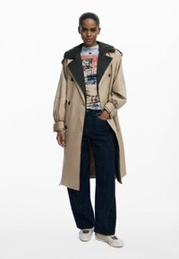 Manteau beige avec col noir, design croisé à double boutonnage. Le mannequin porte un t-shirt graphique multicolore et un pantalon large bleu marine texturé.