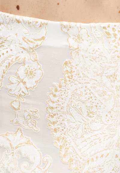 Κοντινή λήψη λευκού υφάσματος με περίπλοκα ανάγλυφα μοτίβα από χρυσά λουλούδια και διακοσμητικά σε φόντο γυμνής επιδερμίδας.
