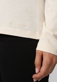 Haut à manches longues crème avec une texture douce, présentant un ourlet cousu et une broderie "MisanGreen" près du bas, porté sur un pantalon noir.