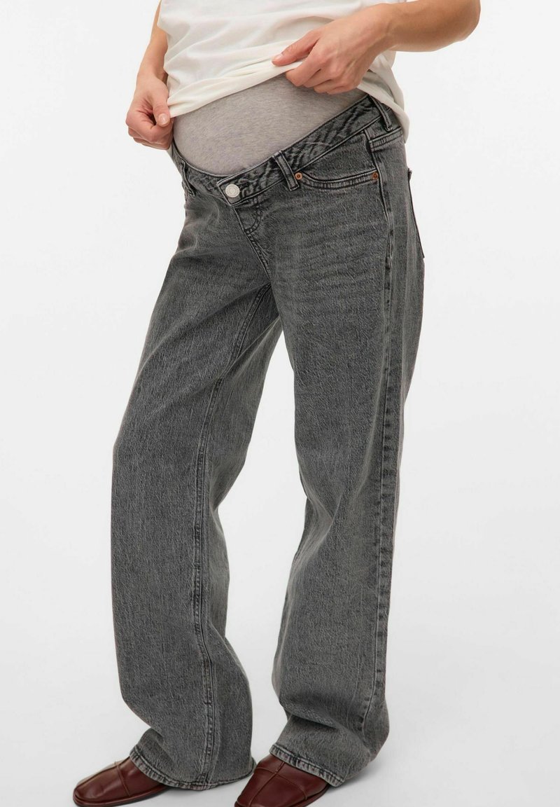 Personne remontant un jean gris ample par-dessus un sous-vêtement taille haute gris clair, portant une chemise blanche et des chaussures marron sur un fond blanc.