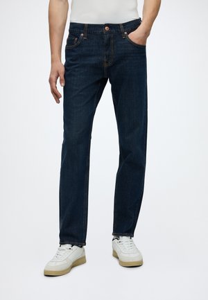 Homme portant un jean droit bleu foncé et des baskets blanches, debout devant un fond uni avec une main dans la poche.