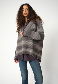 Grau gemusterter Strickpullover mit V-Ausschnitt und geripptem Saum, über einem dunklen Shirt getragen. Besitzt ein kontrastierendes rotes Detail an der Seite.