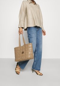 Borsa tote intrecciata beige con due manici, caratterizzata da un logo circolare e una finitura liscia, abbinata a jeans a gamba larga azzurri.