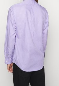 Man i långärmad, lavendelfärgad och vit vertikalt randig skjorta med svarta byxor, vänd bort mot en enkel ljus bakgrund.