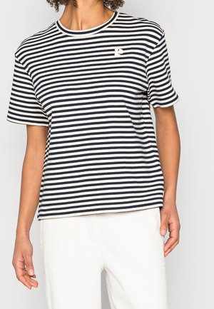 Osoba ubrana w czarno-biały, poziomo paskowany, krótki rękaw i białe spodnie, stojąca na tle jednolitego tła.