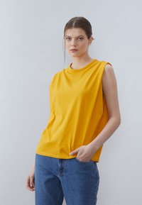 Yellow sleeveless cotton top with a round neckline, relaxed fit, and side slits, paired with blue denim jeans featuring a classic cut.