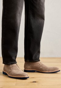 Stivaletti alla caviglia in suede beige con punta arrotondata, design minimalista, rifiniture in pelle marrone chiaro e suola in gomma blu, abbinati a jeans scuri.