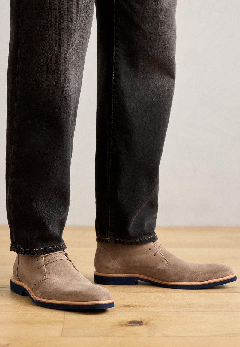 Beige Wildleder Stiefeletten mit runder Zehenpartie, minimalistisches Design, hellbraune Lederverzierungen und blauer Gummisohle, getragen mit dunklen Jeans.