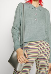 Pull en maille gris clair avec un col et trois boutons décoratifs, associé à un pantalon rayé multicolore. Sac bandoulière argenté à la main.