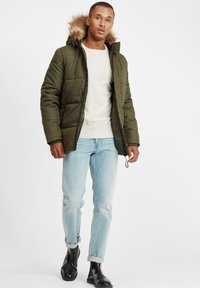 Olivengrønn polstret jakke med fuskepelskant på hetten, glidelås og to sidelommer, iført over en lys grå genser og lyse jeans.