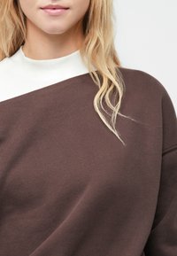 Sudadera marrón con un cuello blanco de alto contraste. Tejido suave, corte oversized y costuras visibles en los hombros.