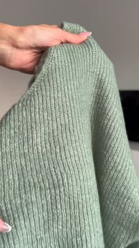 Gaiši zaļš adīts džemperis ar redzamu vertikālu tekstūru un ribu rakstu. Audums izskatās mīksts un mājīgs, to tur roka.