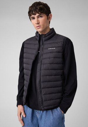 Giovane uomo che indossa un gilet trapuntato nero Guess Jeans sopra una camicia nera a maniche lunghe e jeans blu, in piedi davanti a uno sfondo grigio uniforme.