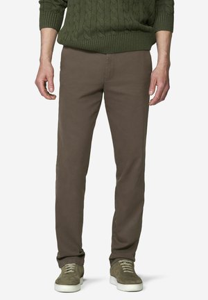 Mann trägt olivgrünen Zopfstrickpullover, braune gerade geschnittene Hose und olivfarbene Velours-Sneaker mit weißen Sohlen, steht vor weißem Hintergrund.