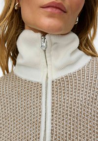 Beež värviga lukuga kampsun, millel on soonikkoes valge kõrge krae; kogu pinnal tekstuurilise koemustriga. Hõbedane lukk läbipaistva tõmbamisdetailiga, sile kangas.