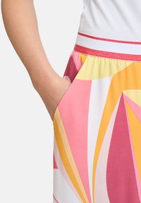Kolorowa, wzorzysta spódnica z odważnymi geometrycznymi wzorami w różowym, żółtym i białym kolorze, z czerwoną, błyszczącą talią i boczną kieszenią.