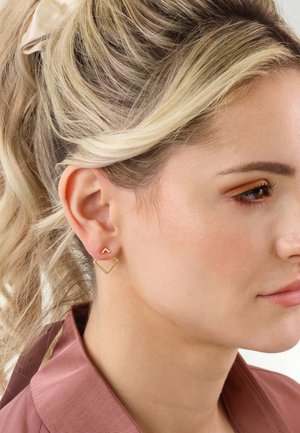 Gold geometric earrings: one with a diamond shape, the other a small triangle, set against a light skin tone, with wavy blonde hair visible.