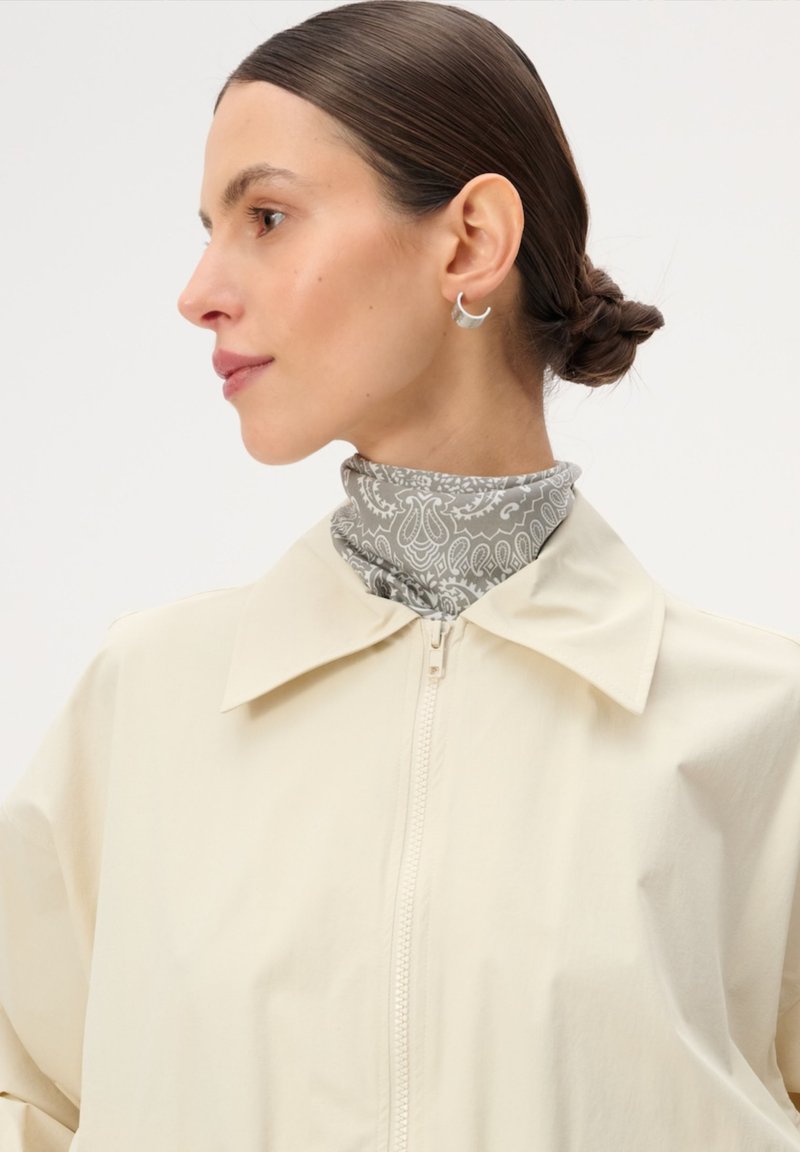 Kobieta z ciemnymi włosami upiętymi w niski kok, nosząca srebrne kolczyki w kształcie półksiężyca, jasny wzorzysty szalik oraz kremową kurtkę na suwak z szerokim kołnierzem.