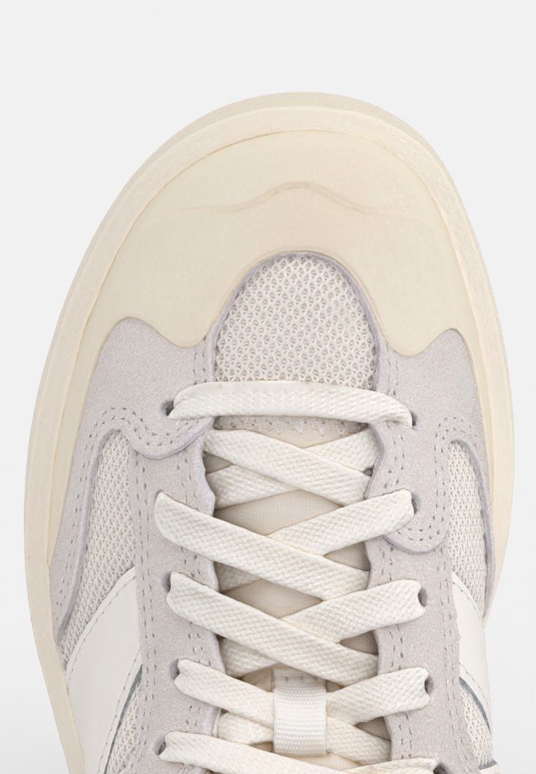Hellgraue Sneaker mit einem Obermaterial aus Wildleder und Mesh, weißen Schnürsenkeln und einer abgerundeten Zehenpartie mit einer tanfarbenen Gummisohle. Akzentuiert mit einem weißen Streifenmuster.