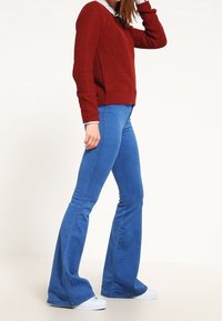 Maglione rosso lavorato a maglia con texture a costine e colletto bianco, abbinato a jeans a zampa d'elefante blu e sneakers bianche, esposto su uno sfondo chiaro.