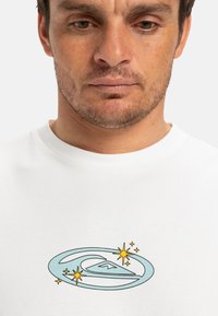 White cotton t-shirt featuring a light blue logo with stars and a wave design at the center. Crew neck and short sleeves.