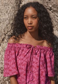 Off-shoulder top i lyserødt stof med blomsterprint, elastisk halsudskæring og bindedetalje med kvaster. Korte, lette ærmer.