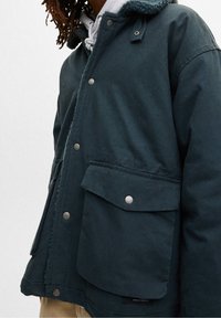 Person som bär en mörkteal jacka med stora framfickor och tryckknappar över en ljusgrå huvtröja.