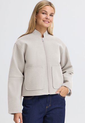 Femme souriante portant une veste zippée gris clair avec deux poches avant et un jean bleu foncé, posant avec une main dans la poche devant un fond uni.