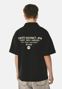 Camisa de manga corta negra con texto blanco en la parte posterior que dice "TOKYO DISTRICT. JPN" junto con detalles de ubicación y caracteres japoneses.