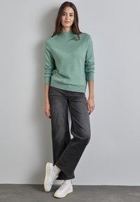 Minzgrüner Strickpullover mit geripptem Hochkragen und strukturiertem Zickzackdesign, kombiniert mit weiten, dunkelgrauen Jeans und weißen Sneakers.