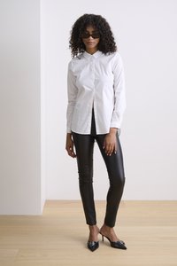 Weißes Hemd mit Knöpfen, schwarze Kunstlederhose, schwarze spitze Absatzschuhe mit dekorativer Hardware und dunkle Sonnenbrille. Glatte Texturen und figurbetontes Design.