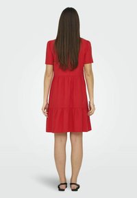 Vestido rojo de manga corta con falda en capas. Textura de algodón suave. Presenta un corpiño ajustado y un acabado acampanado. Llevado con sandalias negras.