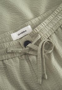 Světlé zelené kraťasy z texturované látky s elastickým pasem, stahovací šňůrkou a černým knoflíkem. Štítek nese nápis "GABBA."