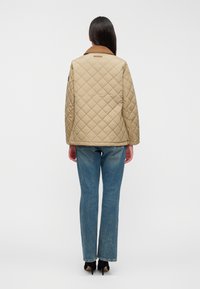 Μπεζ μπουφάν με καπιτονέ σχέδιο και καφέ γιακά, με τετράγωνο μοτίβο. Συνδυασμένο με μπλε τζιν και μαύρα ψηλοτάκουνα, προβολή από πίσω.